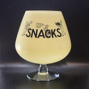 May include: A large, pale yellow glass bowl on a clear pedestal base. The front features black text reading "SNACKS" with illustrations of two chefs and a rooster. A speech bubble above the rooster contains the text "Something to Crow about".