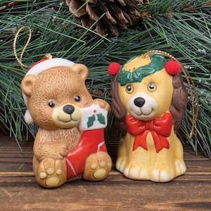 May include: Two ceramic Christmas ornaments: a brown bear wearing a Santa hat and holding a red stocking, and a yellow dog with a red bow and a green wreath. Both ornaments have gold hanging strings and are set against a backdrop of pine needles.