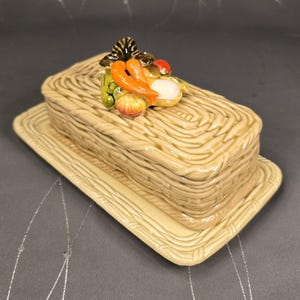May include: A ceramic butter dish with a basket weave design. The lid is topped with a cluster of colorful ceramic vegetables, including carrots, tomatoes, and onions. The dish sits on a matching rectangular tray.
