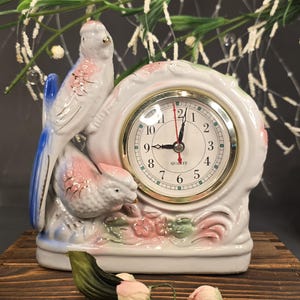 May include: A white ceramic clock featuring two birds with pink and blue accents. The clock face has a gold-colored bezel and displays the time. The clock is decorated with floral details and sits on a wooden surface.