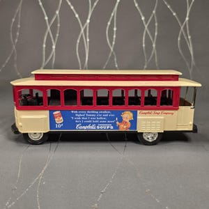May include: A vintage toy trolley car in cream, red, and white, featuring the Campbell's Soup Company logo and a small advertisement. The trolley has black wheels and a detailed interior, with text that reads "Campbells SOUP'S".