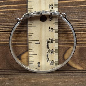 Puede incluir: Brazalete de plata con un acento de cristal rectangular. El brazalete se mide con una regla, mostrando un di&aacute;metro de aproximadamente 7,6 cm. El fondo es una superficie de madera.