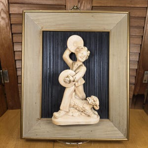 May include: A framed relief sculpture featuring a cream-colored figure of a boy playing a tambourine, with a sheep at his feet. The sculpture is set against a black textured background within a gold and cream-colored frame. The frame is rectangular with a ridged design.