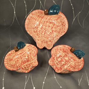 May include: Set of three ceramic apple-shaped bowls. The bowls have a speckled red and cream color with a green leaf and brown stem detail. The bowls are designed with a scalloped edge.