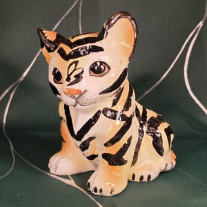 May include: A ceramic tiger figurine with a cream-colored base and black stripes. The tiger has orange ears, nose, and paws. The figurine is sitting with its paws forward, and its eyes are open.