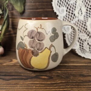 May include: Cream-colored ceramic mug with a brown interior and handle. The mug is decorated with a hand-painted design of grapes, a pear, and leaves in shades of purple, yellow, and green. The mug sits on a wooden surface.