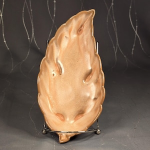 May include: A leaf-shaped ceramic dish in shades of peach and brown, resting on a silver metal stand. The dish has a glossy finish and is designed with detailed leaf-like textures. A decorative piece for serving or display.