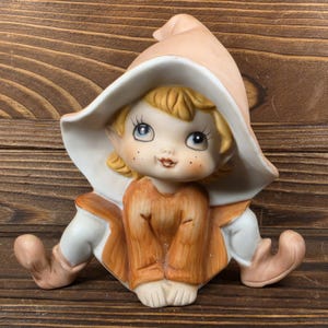 May include: A ceramic figurine of a seated elf with a peach-colored hat and outfit. The elf has blonde hair, blue eyes, and rosy cheeks. The figurine is on a wooden surface.