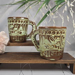 May include: Two ceramic mugs with a brown and green glaze. The mugs have a unique handle design and are decorated with a repeating pattern. The mugs are displayed on a wooden shelf.