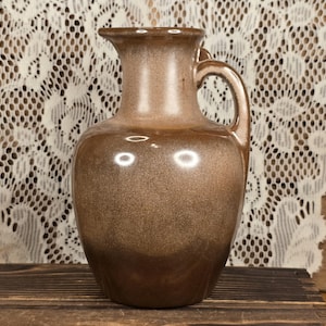 May include: A brown ceramic pitcher with a handle. The pitcher has a wide base that tapers to a narrower neck and a flared rim. The surface has a speckled, metallic finish. The pitcher is sitting on a wooden surface.