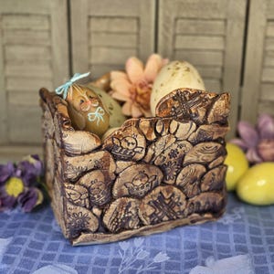 May include: A decorative, brown, stone-textured basket filled with painted Easter eggs and pink flowers. The basket has carved designs of chicks and flowers. One egg is painted with a bunny face and a blue bow.