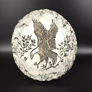 May include: A circular stone-like plaque featuring a relief carving of an eagle with wings spread wide. The eagle is flanked by two leafy branches on either side. The surface has a textured, weathered white and grey finish with dark accents.