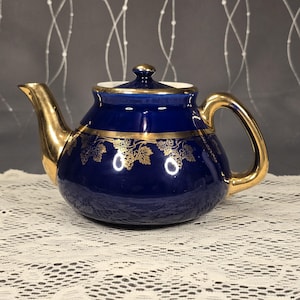 May include: A cobalt blue ceramic teapot with gold accents. The teapot features a gold handle, spout, and lid. A gold grape vine design decorates the body of the teapot. The teapot sits on a white lace tablecloth.