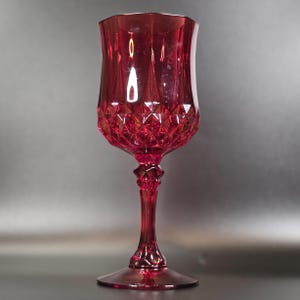 May include: A deep red translucent goblet featuring a faceted diamond-cut pattern on the lower portion of the bowl and a smooth upper rim. The stem is detailed with a decorative geometric node, leading down to a wide, circular base.