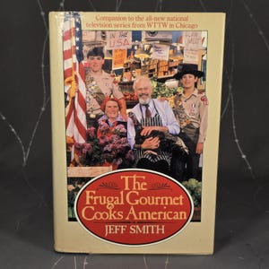 May include: A book cover for "The Frugal Gourmet Cooks American" by Jeff Smith. The cover features a group of people, including two scouts, and the American flag. The title is in a red oval with white text.