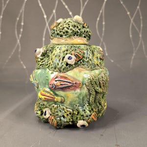 May include: A ceramic jar with a lid, featuring a marine theme. The jar is primarily green with textured coral-like patterns and colorful fish accents. The lid has a similar coral design. The jar is approximately 6 inches tall.
