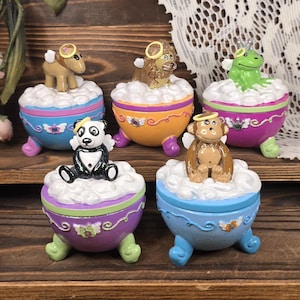 May include: A collection of whimsical, decorative trinket boxes. Each box features a different animal figurine, including a panda, monkey, lion, frog, and a sheep, all with angel wings and halos, atop a cloud-like design. The boxes are painted in bright colors.