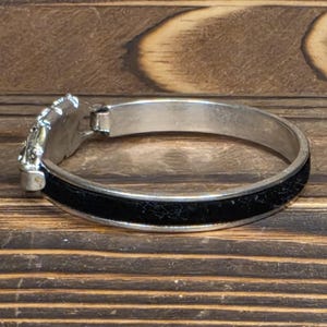 Puede incluir: Pulsera articulada plateada con una banda negra. La pulsera presenta un cierre decorativo plateado con un dise&ntilde;o ornamentado. La banda negra parece texturizada. La pulsera se exhibe sobre una superficie de madera.