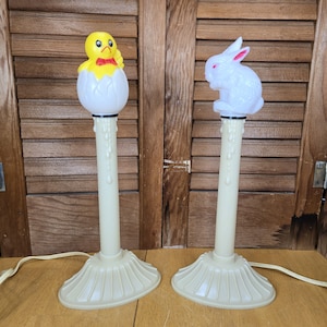 May include: Two Easter-themed candle lamps. One lamp features a yellow chick emerging from a white egg, wearing a red bow tie. The other lamp has a white bunny with pink ears. Both lamps have a cream-colored base and stem, with a dripping wax design.