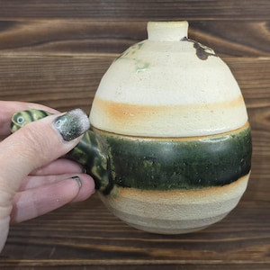May include: A ceramic lidded pot with a handle. The pot has a rounded shape with a cream-colored top and a dark green band around the middle. The handle is green with a swirl design. The pot is held by a person.