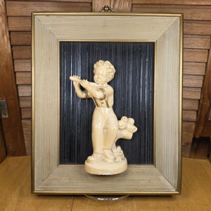 May include: A framed vintage relief sculpture of a boy playing a flute. The sculpture is cream-colored and set against a black background within a gold-trimmed frame. The frame has a cream-colored, ribbed border.