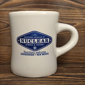 May include: White ceramic mug with a curved handle. The mug features a blue and white logo for "The National Museum of Nuclear Science & History" with the text "Reactions welcome. Albuquerque, New Mexico."