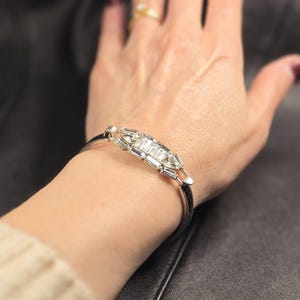 Puede incluir: Una pulsera plateada con un dise&ntilde;o rectangular que presenta m&uacute;ltiples piedras rectangulares transparentes. La pulsera se lleva en la mu&ntilde;eca, con un anillo de oro visible en un dedo. El fondo es una superficie oscura y texturizada.