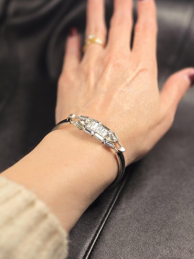 Puede incluir: Una pulsera plateada con un dise&ntilde;o rectangular que presenta m&uacute;ltiples piedras rectangulares transparentes. La pulsera se lleva en la mu&ntilde;eca, con un anillo de oro visible en un dedo. El fondo es una superficie oscura y texturizada.
