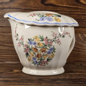 May include: A white ceramic pitcher with a lid, adorned with floral patterns in shades of pink, yellow, blue, and green. The pitcher has a handle and a scalloped rim with a blue trim. The floral design includes butterflies.