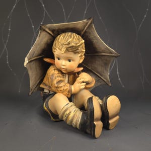 May include: A ceramic figurine of a young child sitting under a brown umbrella. The child wears a tan jacket, black shorts, and black boots. The figurine has a detailed face and is a decorative item.