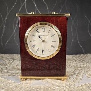 May include: A rectangular Howard Miller clock with a cherry wood finish and gold-tone accents. The clock face is white with black Roman numerals and hands. The clock is a classic decorative piece.