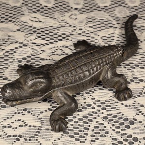 May include: A dark, cast-iron alligator figurine with detailed scales and legs. The alligator is lying down with its tail curved upwards. The figurine is on a white lace surface.