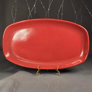 May include: A vibrant red, oval-shaped ceramic platter. The platter has a slightly speckled texture and rounded edges. It is displayed on a decorative gold-colored metal stand, showcasing its shape and color. The background is a dark gray.