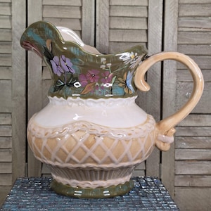 May include: A ceramic pitcher featuring a scalloped rim and a handle. The upper section is olive green with painted purple and white flowers. The lower section is cream-colored with a raised, tan-colored lattice pattern and a dark green base.