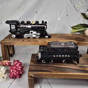 May include: Two black ceramic train figurines, one marked "773" and the other "Lionel", sit on wooden shelves. The train engines have white and gold accents. The background is a light gray with floral accents.