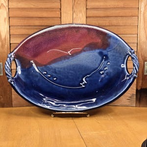 May include: A handmade ceramic serving platter with a unique design. The oval-shaped dish features a gradient of colors, transitioning from deep red to blue. It has rope-like handles and a white abstract design. The platter is displayed on a metal stand.