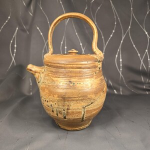 May include: A handcrafted ceramic teapot with a lid and a curved handle. The teapot is primarily brown with lighter and darker brown bands and drips of green glaze. The spout is on the left side. The overall shape is rounded.