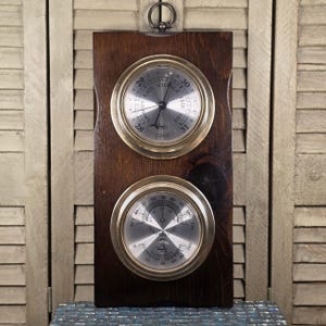 May include: A wall-mounted weather station on a dark brown wooden board featuring two circular brass-rimmed gauges. The top gauge is a barometer with a silver face labeled Rain, Change, and Fair. The bottom gauge is a thermometer and hygrometer.