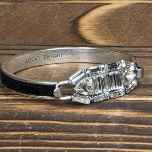Puede incluir: Brazalete de tono plateado con una banda negra y un dise&ntilde;o rectangular decorativo de cristal. El brazalete est&aacute; marcado "LUCKY BRAND" en el interior. Los cristales son transparentes y facetados, a&ntilde;adiendo brillo.
