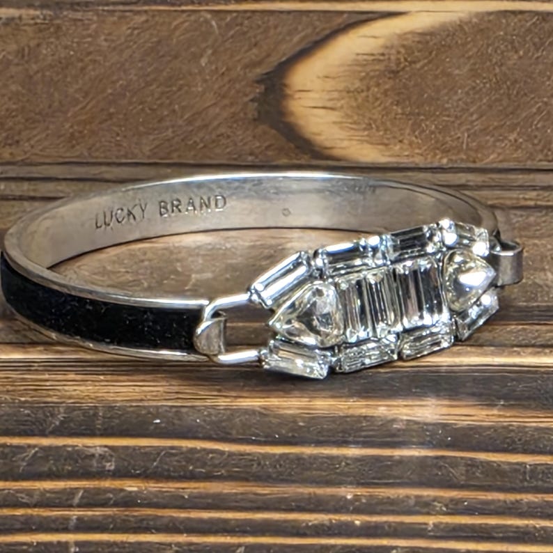 Puede incluir: Brazalete de tono plateado con una banda negra y un dise&ntilde;o rectangular decorativo de cristal. El brazalete est&aacute; marcado "LUCKY BRAND" en el interior. Los cristales son transparentes y facetados, a&ntilde;adiendo brillo.