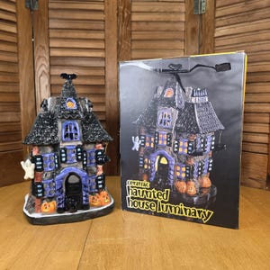 May include: A ceramic haunted house luminary with a black roof, purple accents, and jack-o'-lanterns. A ghost figure hangs from the side. The box next to it reads "ceramic haunted house luminary".