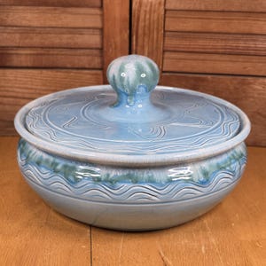 May include: A light blue ceramic lidded bowl with a decorative wave pattern around the base and lid. The lid has a rounded handle and a matching wave design. The bowl is on a wooden surface.