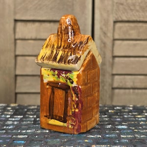 May include: A ceramic house-shaped container with a thatched roof lid. The container is brick-colored with a brown window and a yellow and green painted design. The roof is tan and brown.