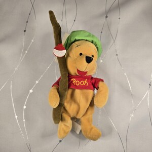 May include: A plush Winnie the Pooh toy wearing a green hat and a red shirt with the name Pooh embroidered in yellow. The character is holding a brown plush fishing rod with a red and white bobber attached to the side of the rod.
