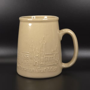 May include: A beige ceramic mug featuring a raised, embossed relief design of the Smithsonian Institution building on its exterior. The text Smithsonian Institution is clearly visible in a script font at the base of the architectural illustration.