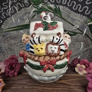 May include: A ceramic Noah's Ark cookie jar. The jar features a white base with a decorative boat design. The ark is adorned with animal faces, including a lion, zebra, and bear. A rooster sits atop the ark. A red bow is on the front.