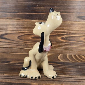 May include: A ceramic figurine of a cartoon dog in a seated position, with its head tilted back and mouth open. The dog has a cream-colored body, black ears, and black spots on its feet. The figurine is on a wooden surface.