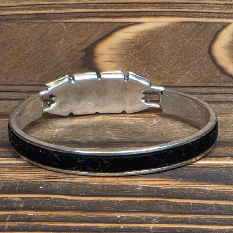 Puede incluir: Una pulsera de brazalete articulada plateada con una banda negra texturizada. La pulsera presenta una pieza central rectangular plateada con peque&ntilde;as incrustaciones rectangulares. La pulsera se exhibe sobre una superficie de madera.