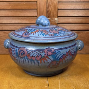 May include: A round blue ceramic lidded casserole dish featuring a hand-painted pattern of pink swirls and dark blue leaf-like motifs. The lid is topped with a small, spiraled handle, and the sides include two small, curled handles on each side.