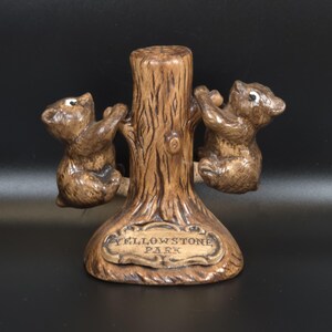 May include: A brown ceramic souvenir figurine featuring two small bears climbing a central tree trunk. A plaque at the base is embossed with the text YELLOWSTONE PARK. The bears have white painted eyes and textured fur details on the tree bark.